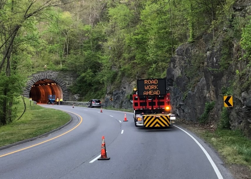 A truck with a sign that says "Road Work Ahead" beside a line of orange cones approaching a tunnel.