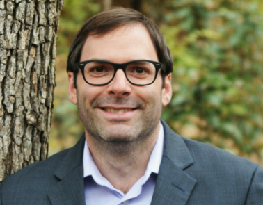 A portrait-style photo of a person standing beside a tree. The individual has short, brown hair and glasses.