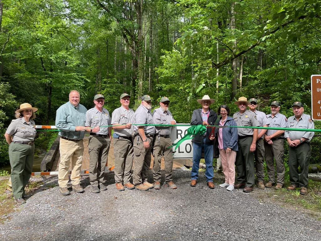 Twelve individuals standing behind a green, commemorative ribbon, on a gravel road surrounded by green trees.