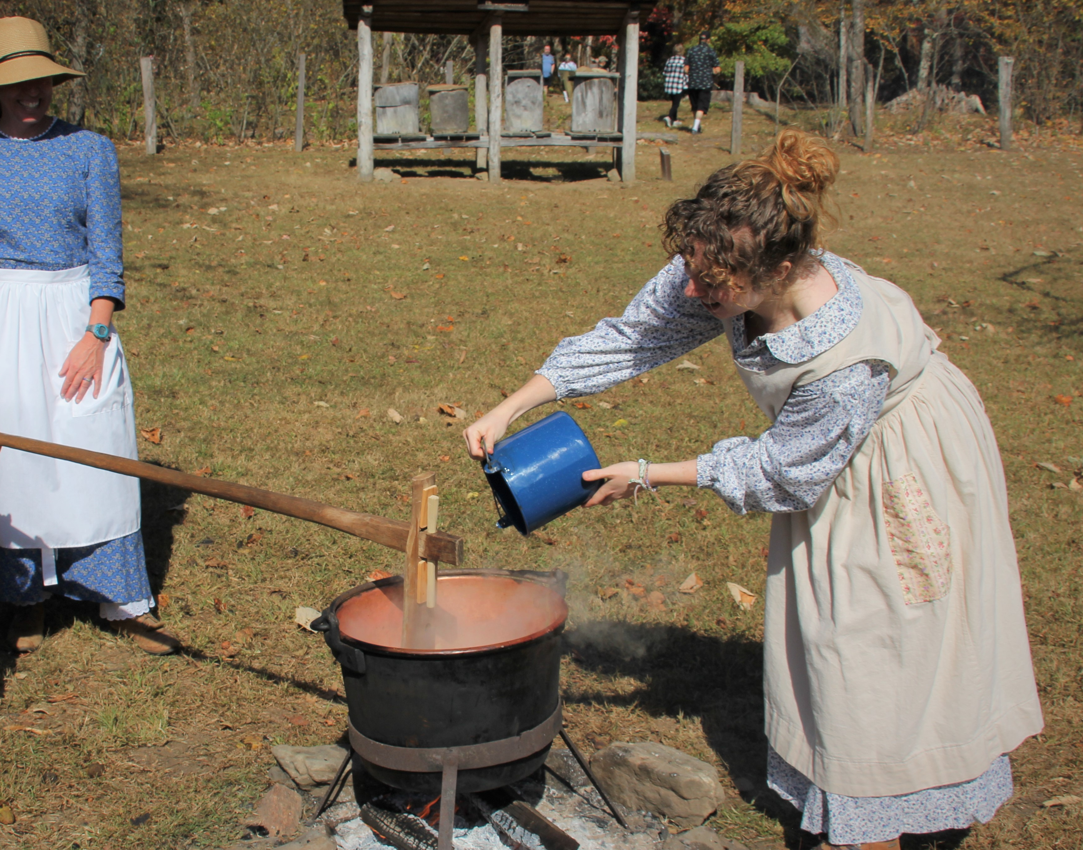 A woman in 1900s dress pouring water into a steaming bucket while another woman stands by.