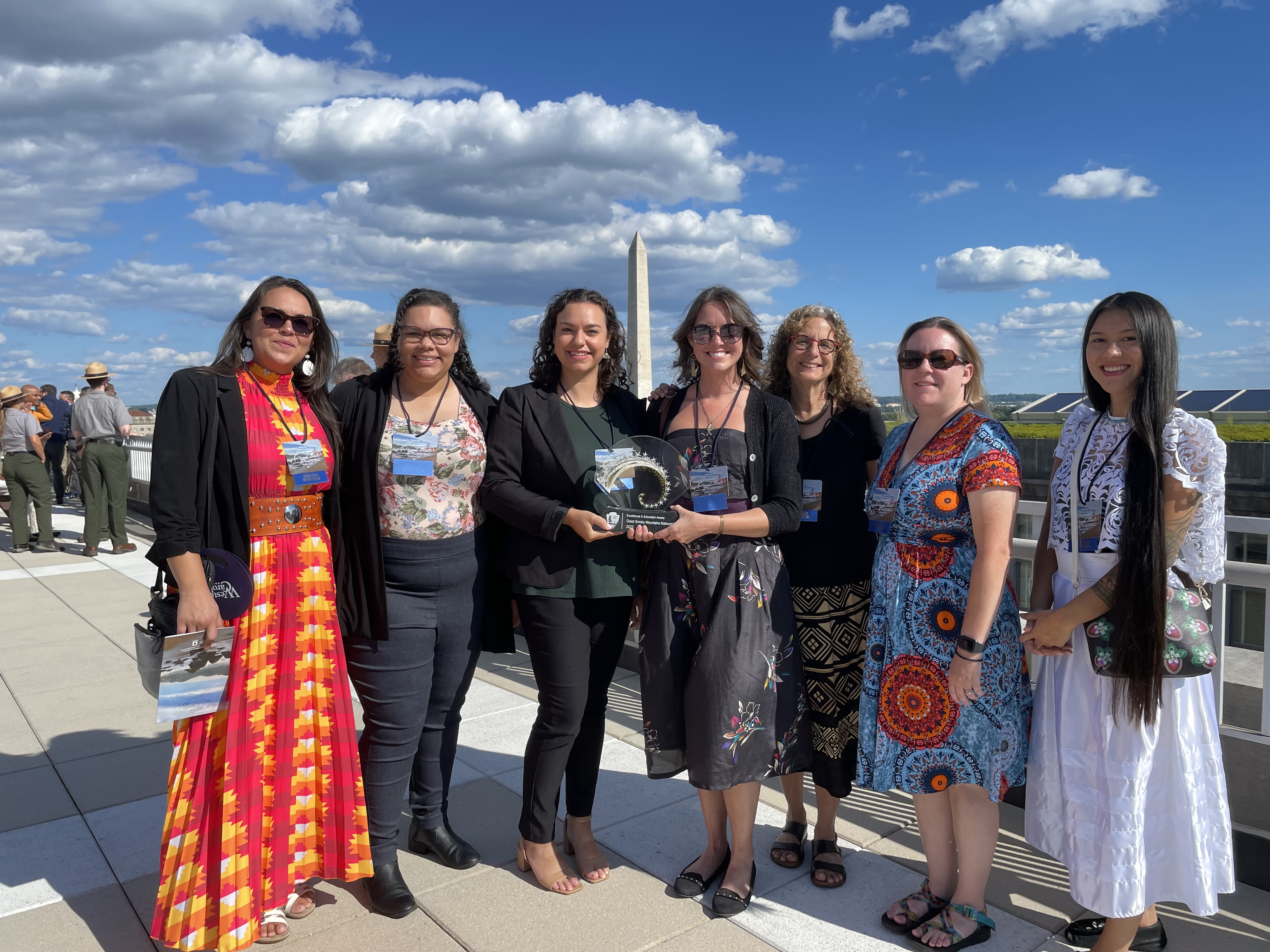 2023 Excellence In Education Awardees Seven people smiling in a row with the Washington Memorial in the background. The two center people hold a circular award. Park rangers and other people are in the left frame background.