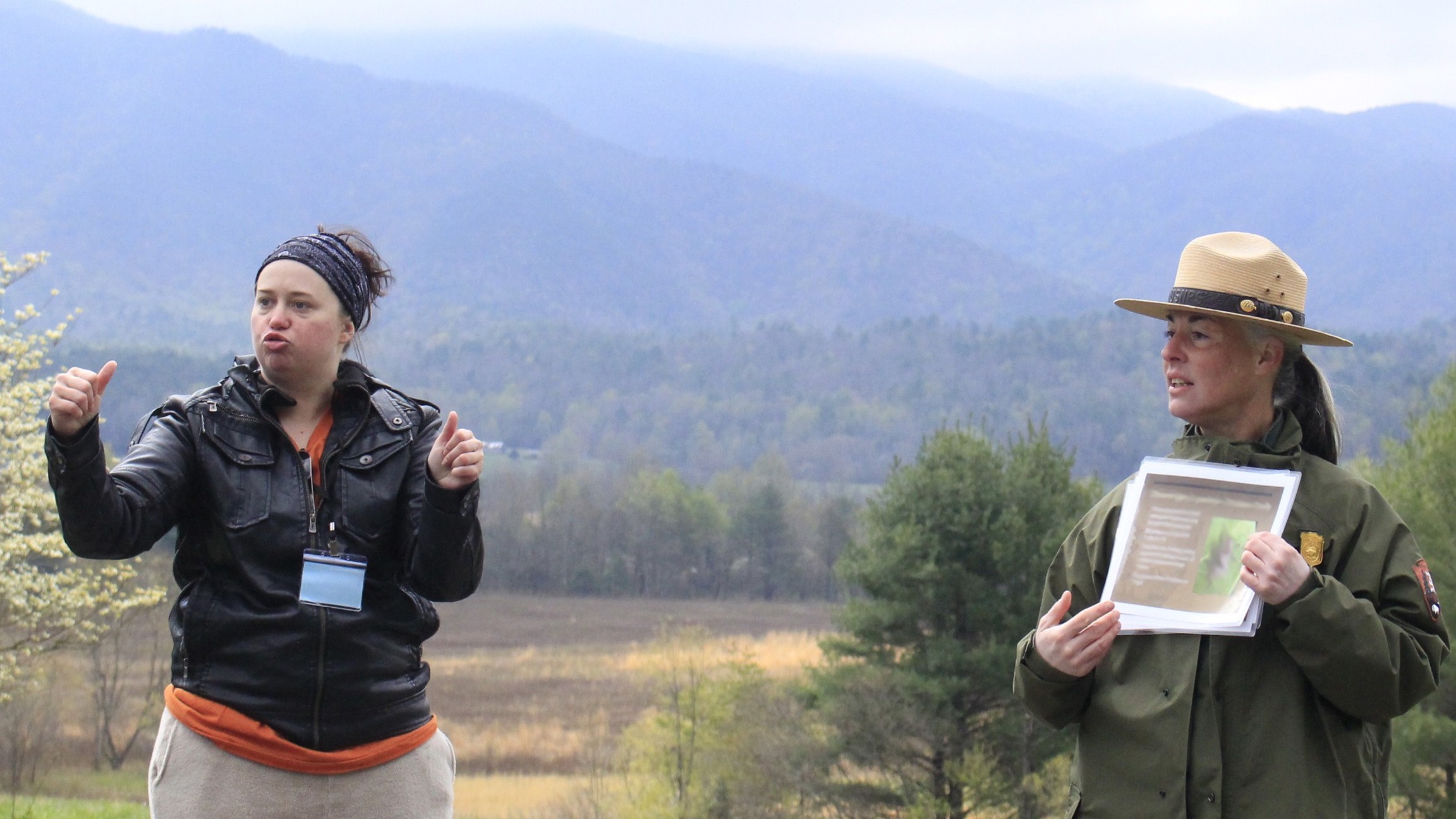 An ASL interpreter stands beside a ranger giving a program with a scenic mountain background.