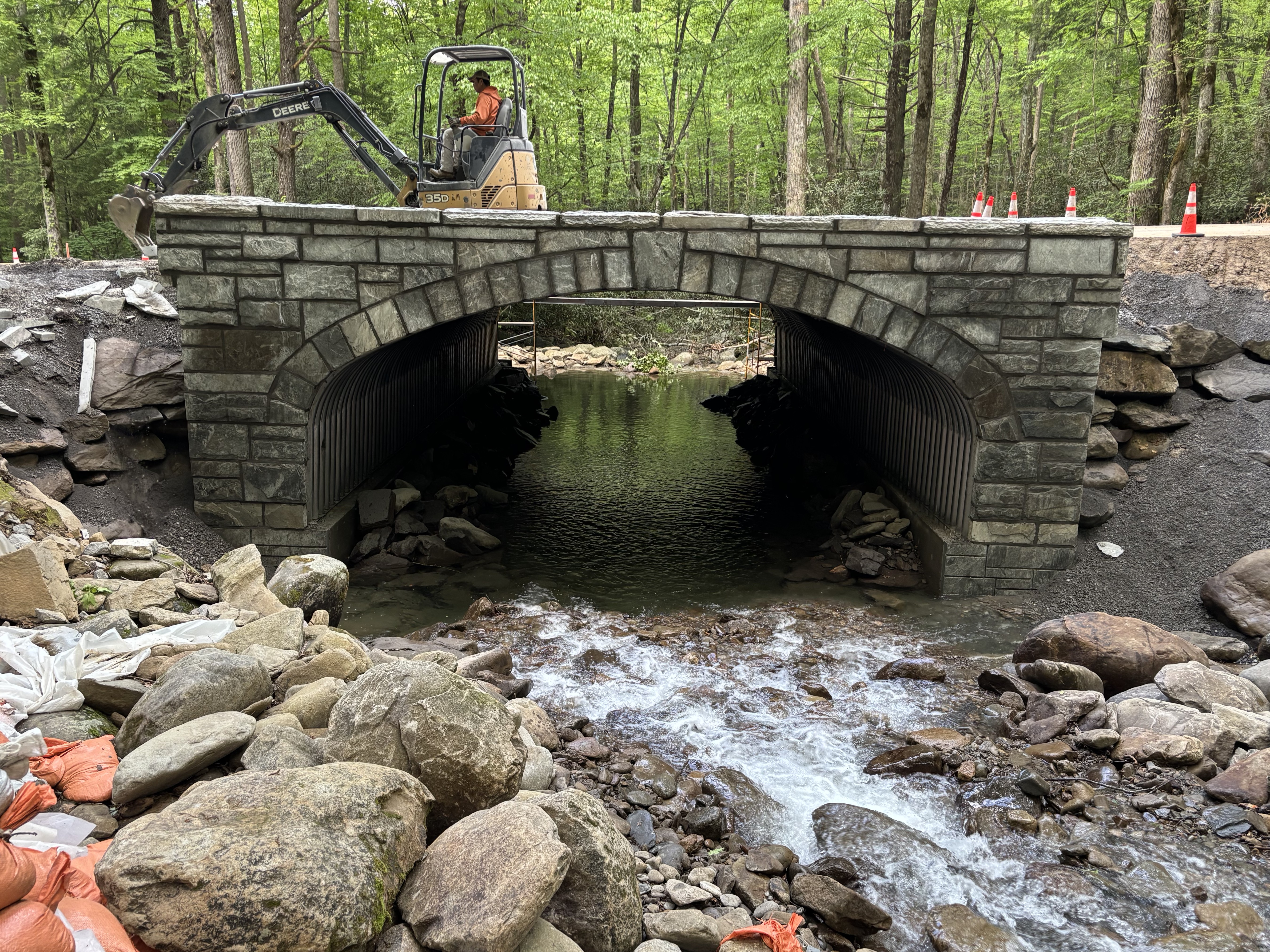 A stone culvert on a roadway being worked on by a crew. A rocky river flows through the culvert.