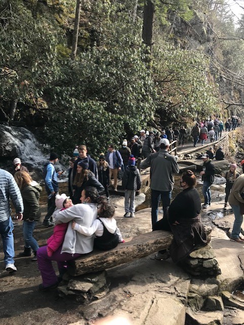 Large crowd of people at waterfall and walking on trail near waterfall in the winter.