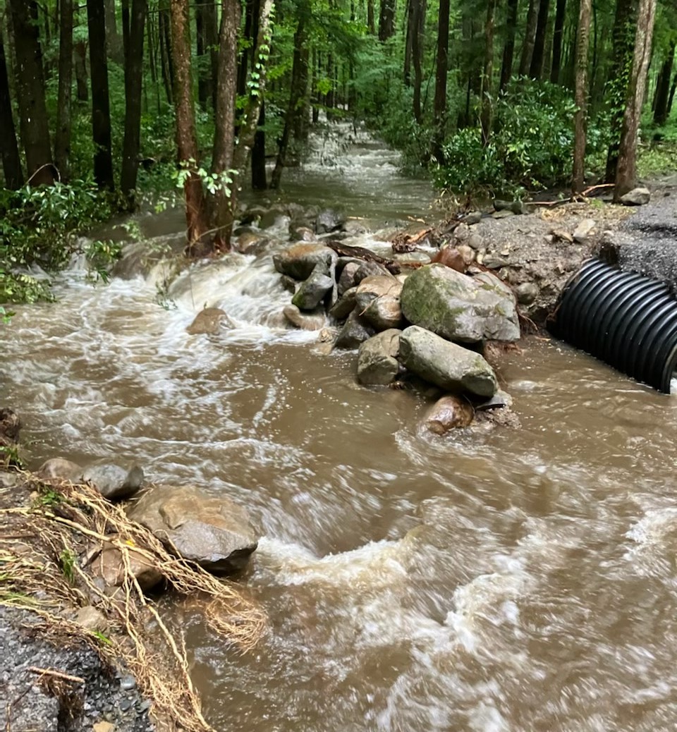 7.21.22 Porters Creek Road flooding Rushing water passing through a wooded area and between two broken pieces of road surrounded by rocks, a culvert, and gravel/grass debris