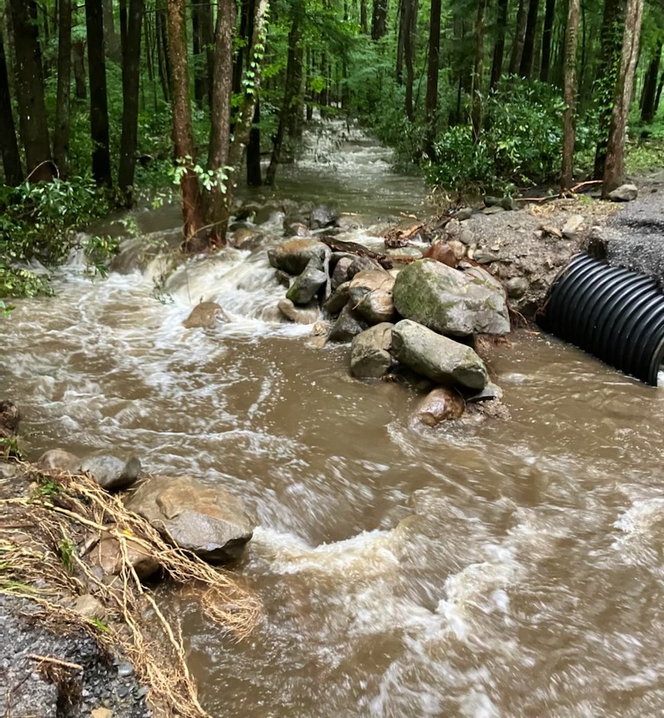 Rushing water passing through a wooded area and between two broken pieces of road surrounded by rocks, a culvert, and gravel/grass debris