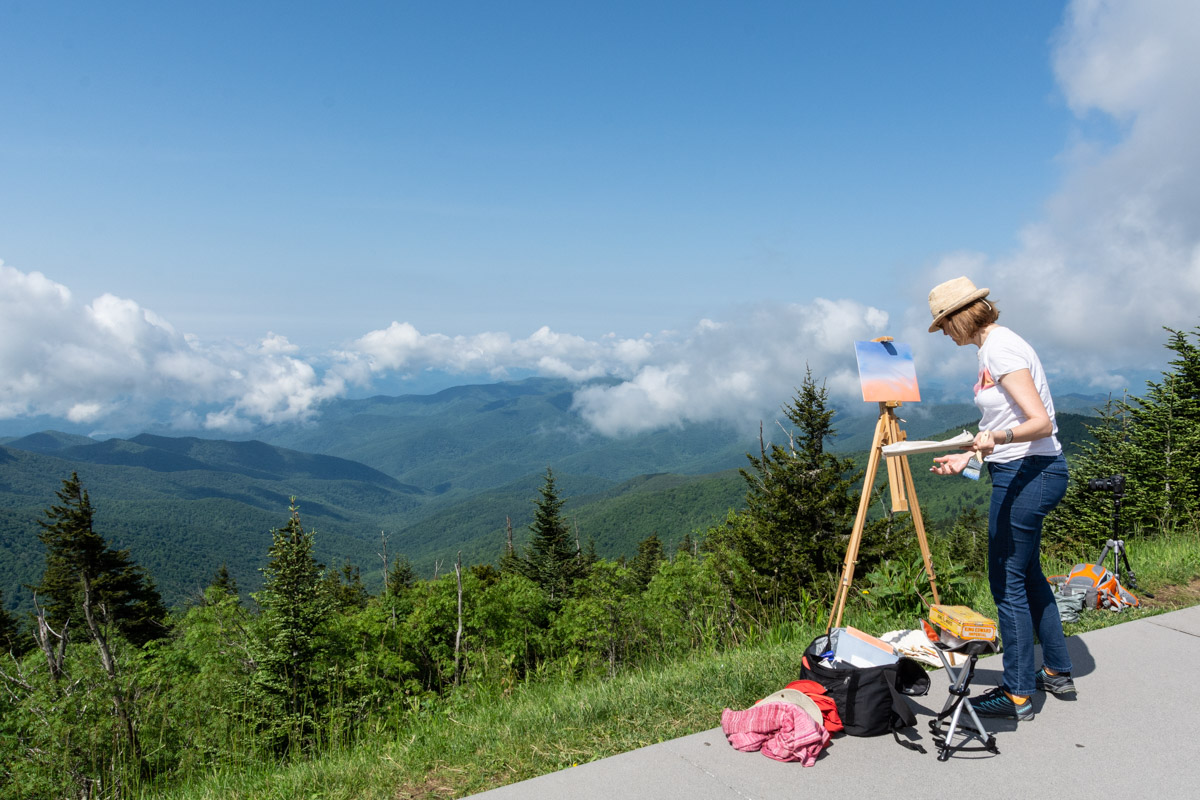 A person standing on a sidewalk painting on a canvas propped on an easel in front of a mountain view.