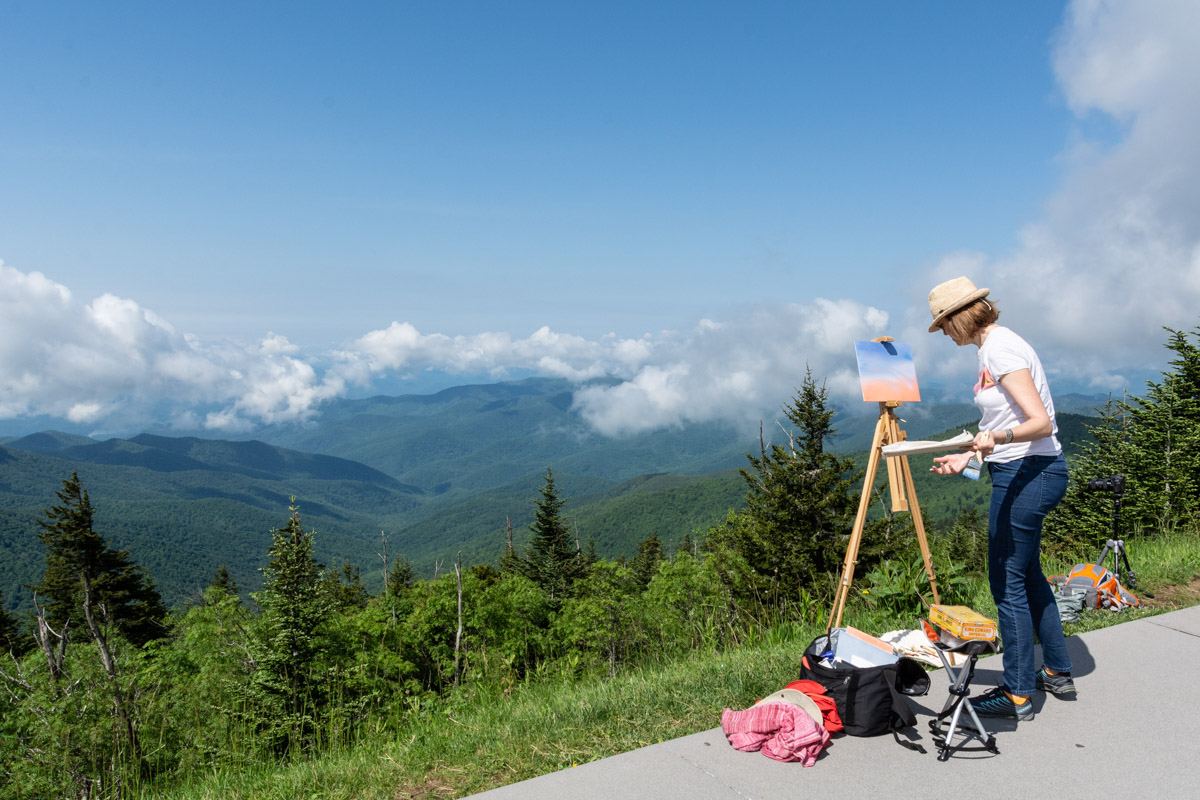 Plein Air Artists at Clingmans Dome on 6.12.18_Photo by Kristina Plaas An artist wearing jeans, a t-shirt, and a hat, stands at an easel painting a mountain view. Green trees and grass are in the foreground. Dispersed clouds cast shadows on the rolling mountains.