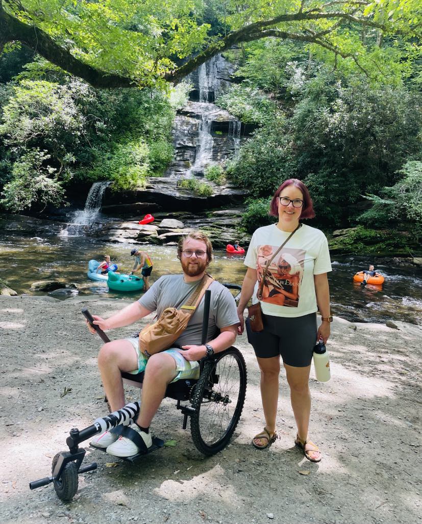 A person in an off-road wheelchair is next to a person standing. They are in front of a waterfall and creek.