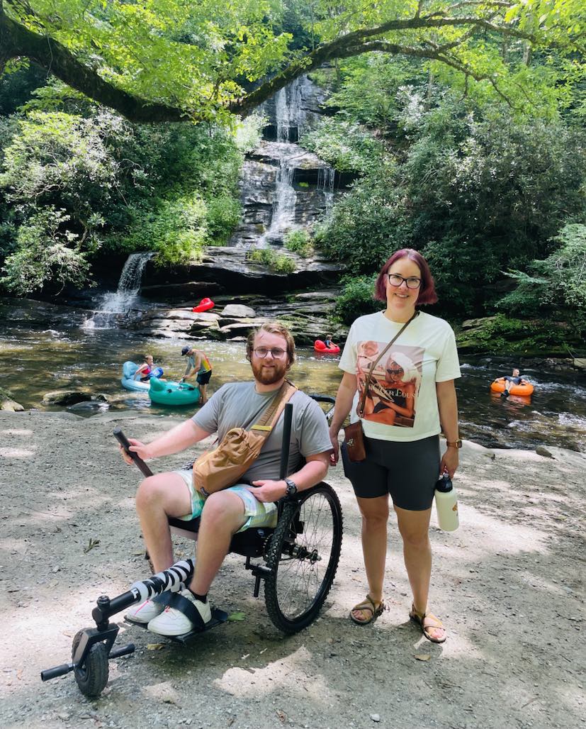 Deep Creek Adaptive program A person in an off-road wheelchair is next to a person standing. They are in front of a waterfall and creek.