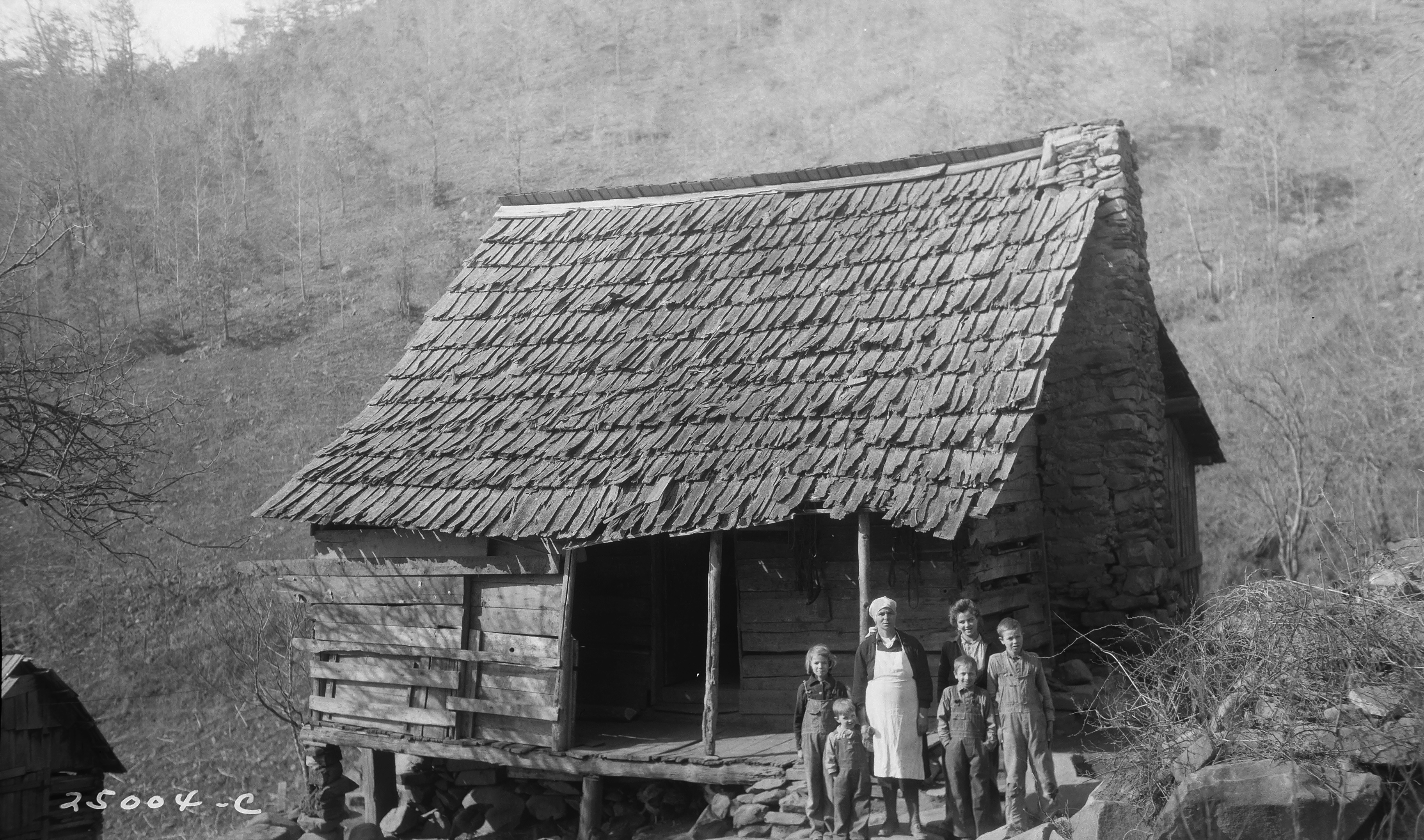 Black and white historic photo with a family of six people standing in front of a wooden home with a stone chimney.
