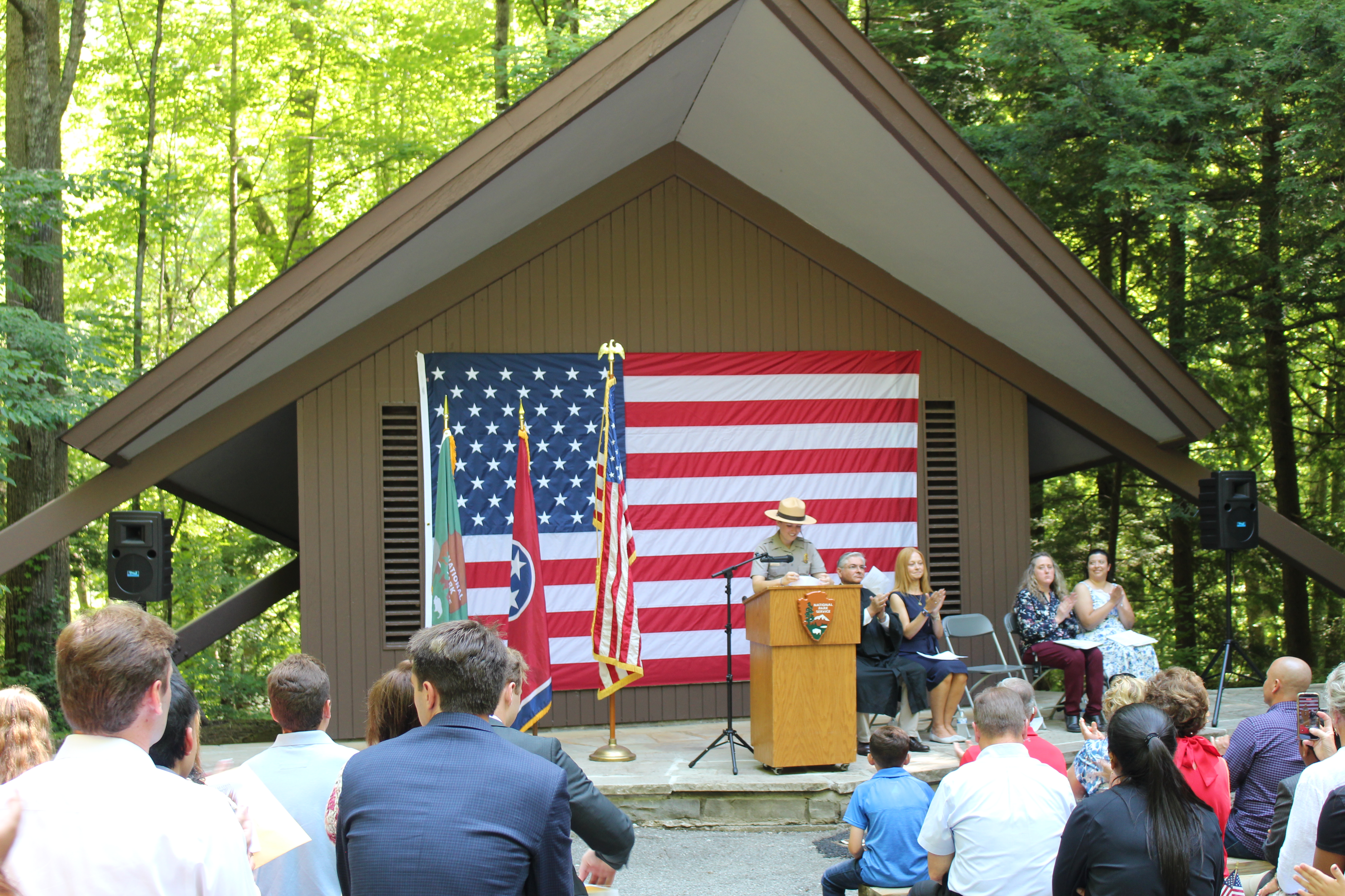 A park ranger speaks to an applauding crowd in front of a large American flag.