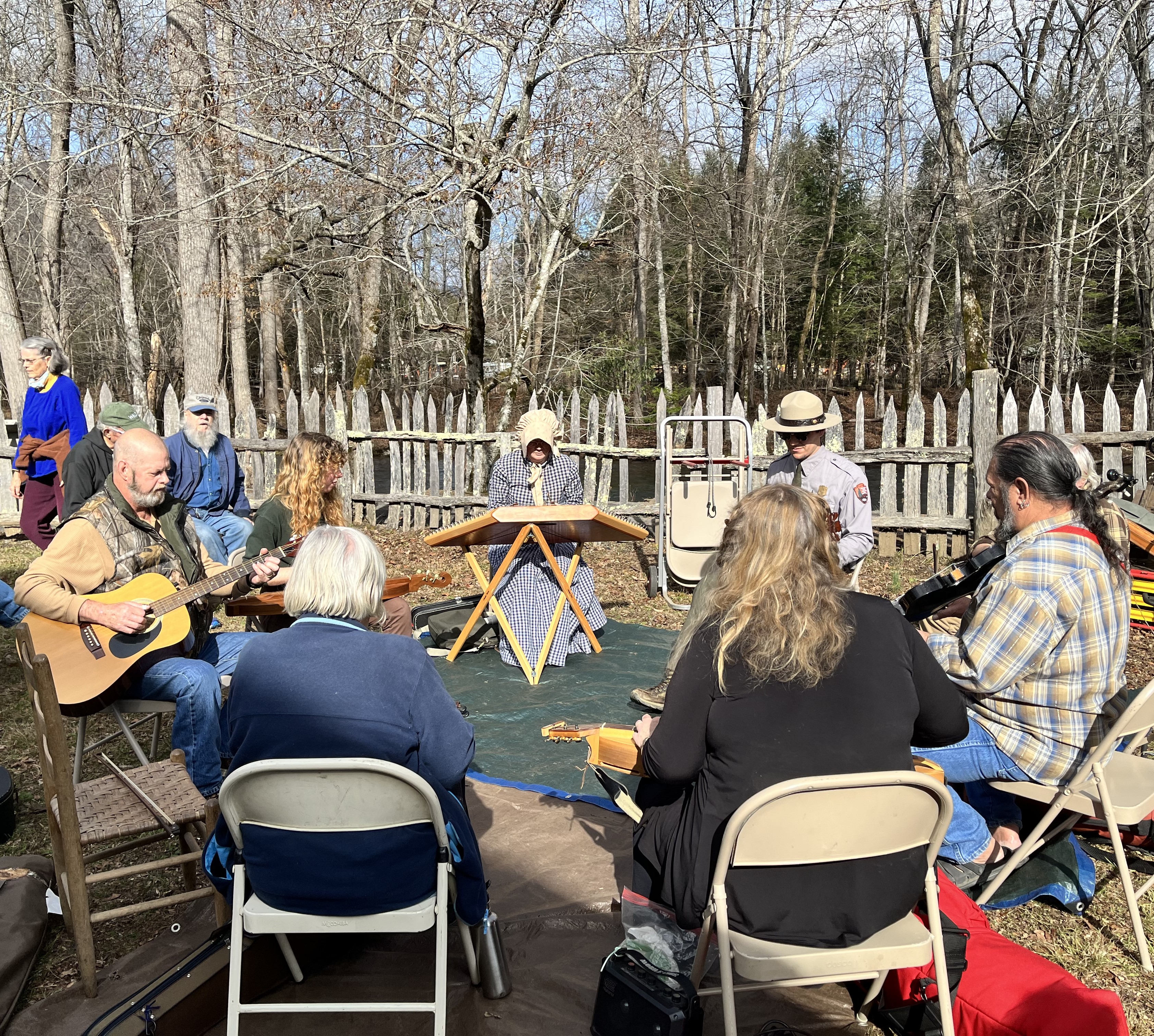 Jam session With trees and a wood fence in the background, a group of people sit in a circle and play guitars, violins, and dulcimers