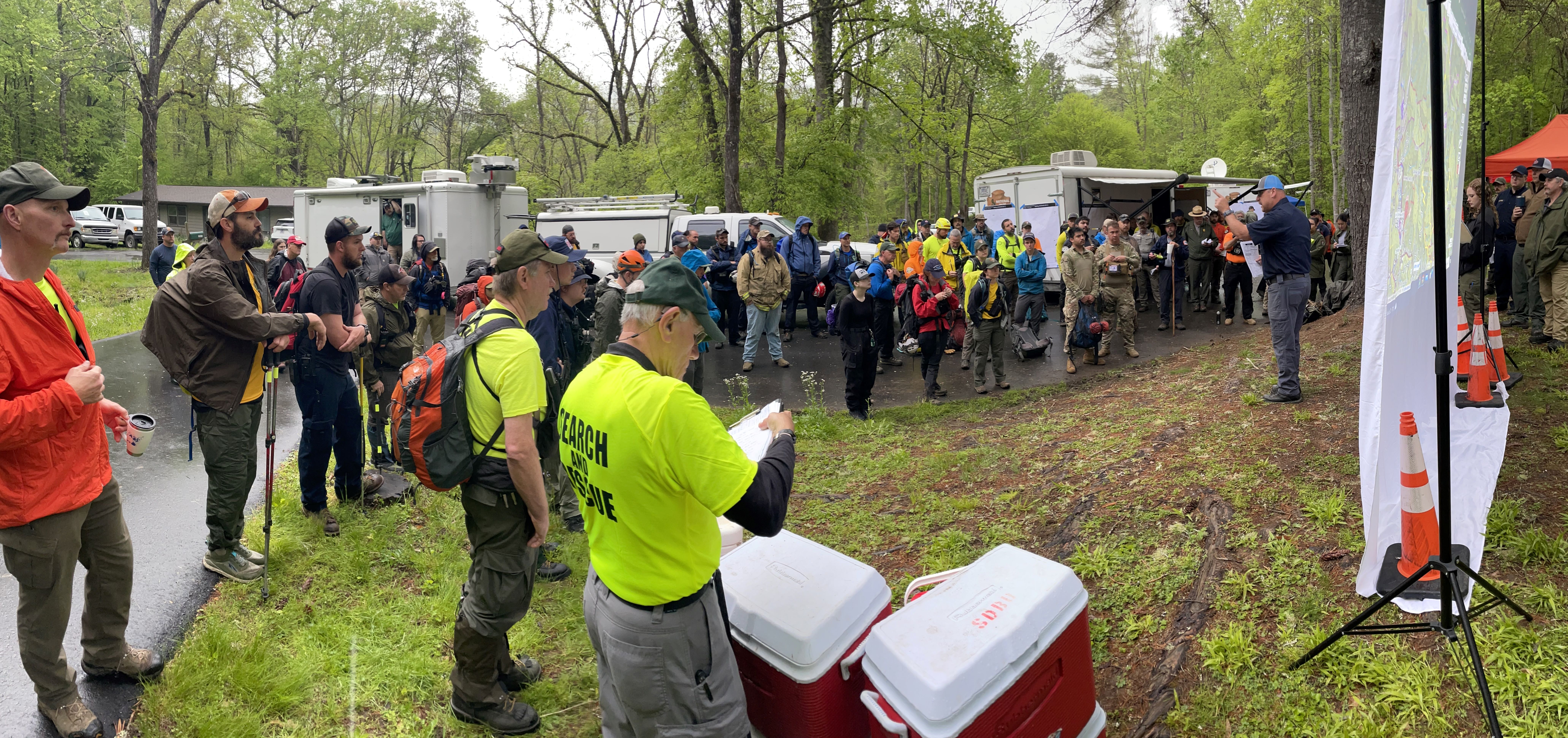 search team members gather for briefing under trees