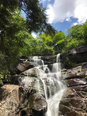 View from below of water cascading over rocks with trees and blue sky with white clouds in the background.