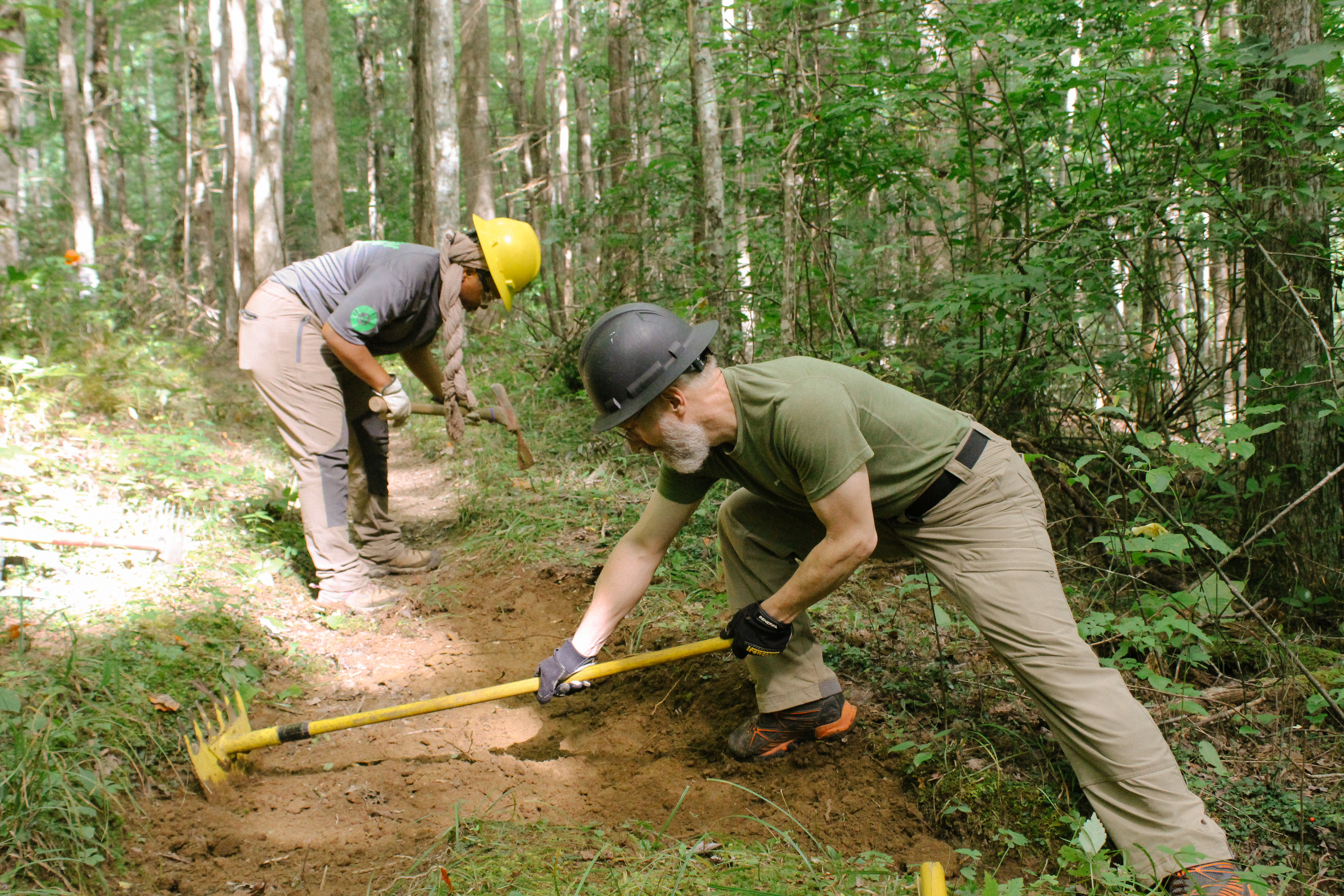 Trails volunteers using hand tools to improve a trail Two people wearing yellow hard hats use hand tools to smooth a brown dirt trail