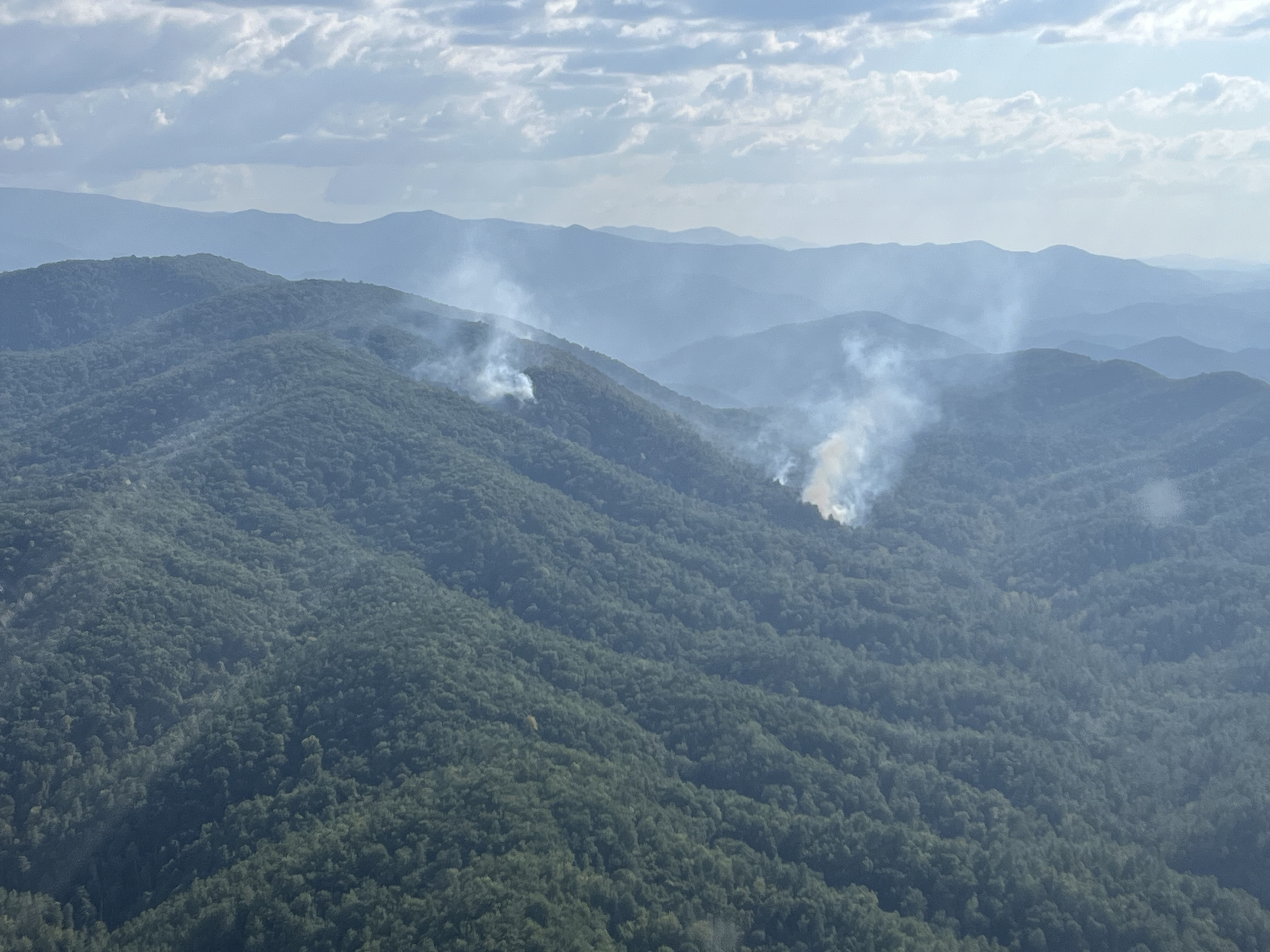 View of Flint Gap Fire 9.16.2024 Two plumes of smoke rising from between rolling mountain ridges on a partly cloudy day.