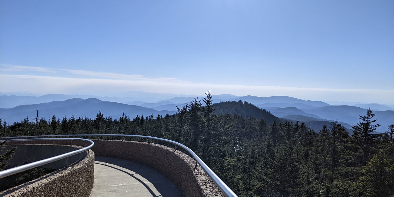 Rolling mountains in the distance. A paved path with metal railing in foreground overlooking nearby evergreen trees.