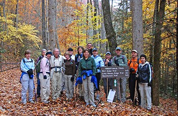 A group of hikers posing beside a wooden trail sign. Colorful fall foliage surrounds the group.