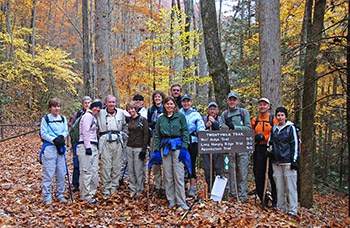 A group of hikers posing beside a wooden trail sign. Colorful fall foliage surrounds the group. A group of hikers posing beside a wooden trail sign. Colorful fall foliage surrounds the group.