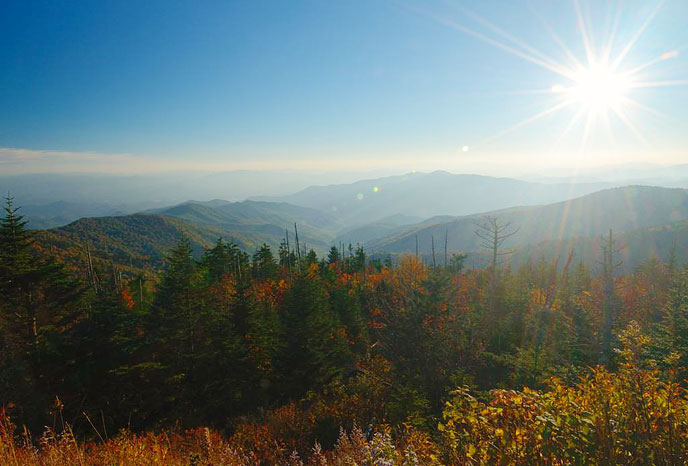 The late afternoon sun is low on the horizon in a blue sky accenting colorful fall trees in the foreground and ridge after ridge of mountains stretching off into the distance.