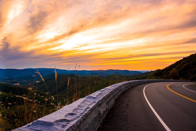 An orange and yellow sunset above rolling mountains in the background of a curve in a road.