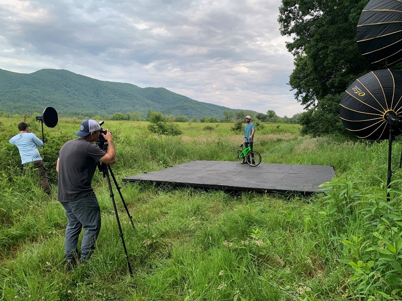 A film crew photographs a person a platform with a bike in a grassy field with mountains behind.