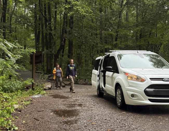Two visitors walking toward a white shuttle van parked beside a trailhead.
