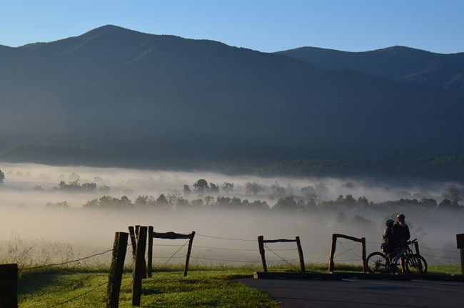 Two people standing by their bikes near a fence with a foggy field and mountains in the background.