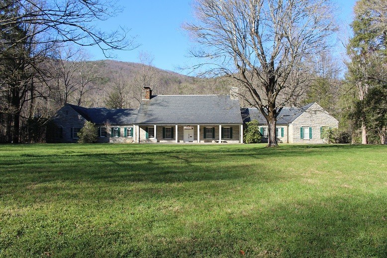 A large field with a gray stone building and green shutters. A large tree sits in front of it and a mountain behind it.