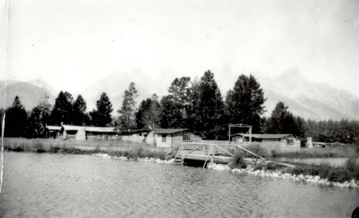 Several log buildings are viewed across a natural swimming pool, surrounded by pine trees, with snow-capped peaks beyond.