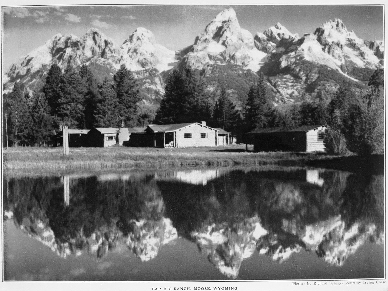 Several log buildings are viewed across a reflecting pool, surrounded by pine trees, with snow-capped peaks beyond.
