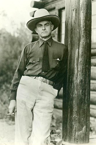 A man in a cowboy hat, with shirt and tie, leans against the post of a log home.