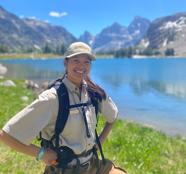 A woman smiling with a lake, mountains, and blue sky in the background.