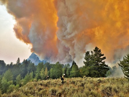 Firefighters wet down a field of sagebrush as a fire puts up a column of smoke in the background.