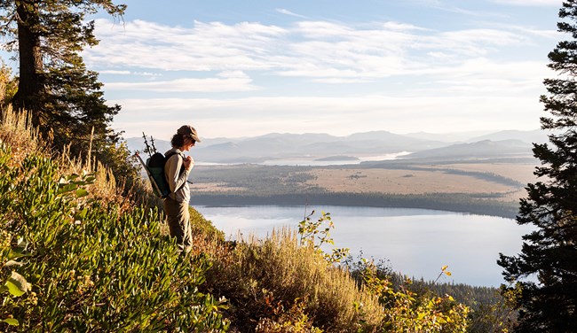 A woman stands on a mountainside trail looking down on a lake in the valley below.