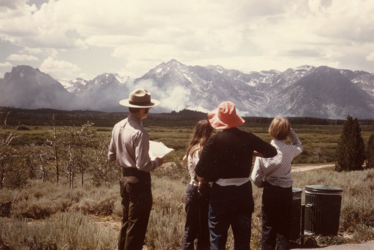 A park ranger talks to three visitors about a fire visible on distant peaks.