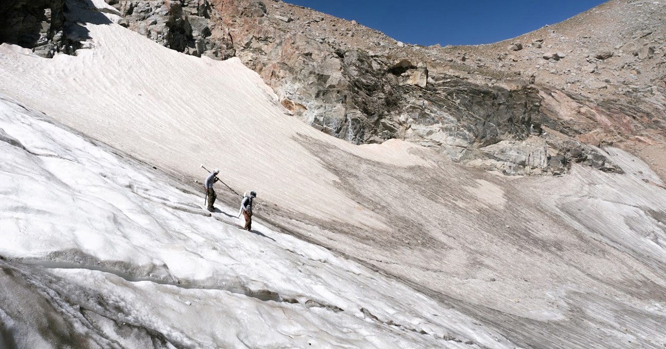 Two scientists walk on the slanted ice of a glacier.
