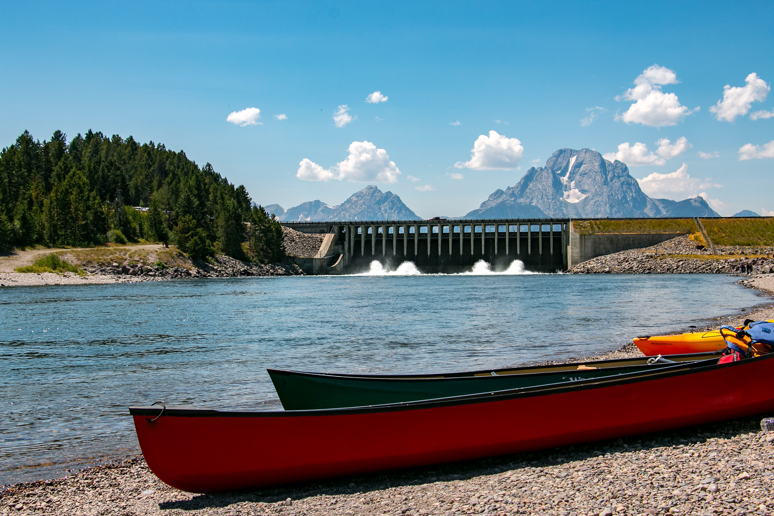 Canoes sit along the shore of the Snake River with the Jackson Lake Dam and Teton Range in the background