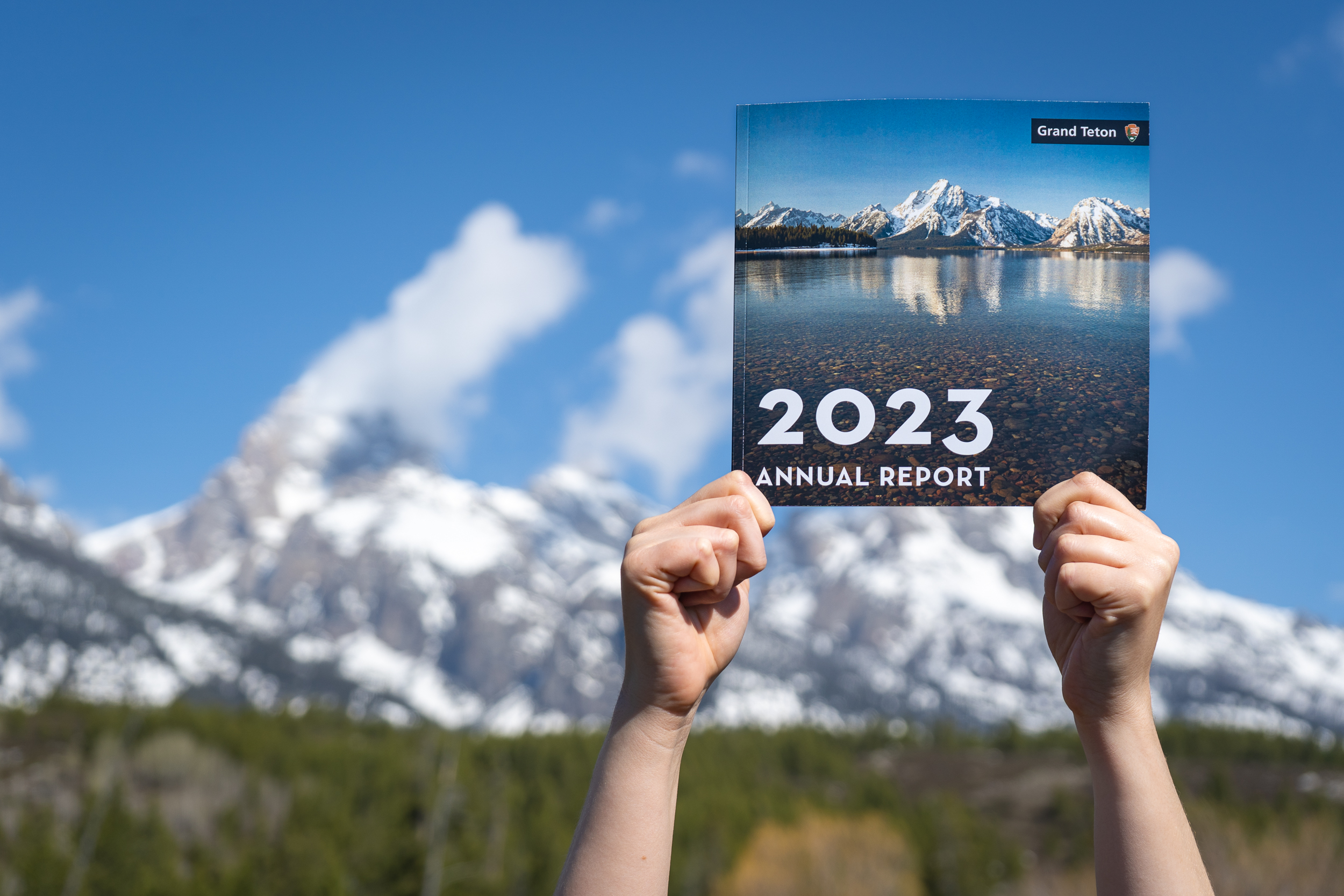 Hands holding the Grand Teton 2023 Annual report in front of the Teton Range and blue skies above.