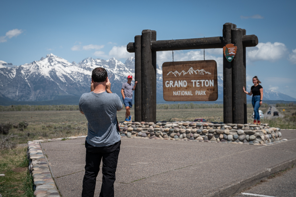 Visitors take picture and Grand Teton National Park entrance sign with the Teton range in the background