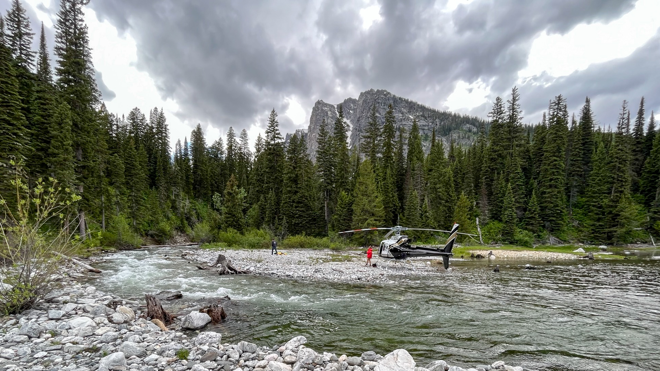 A man and woman stand next to a helicopter parked on a rocky lakeshore with forest and mountains behind