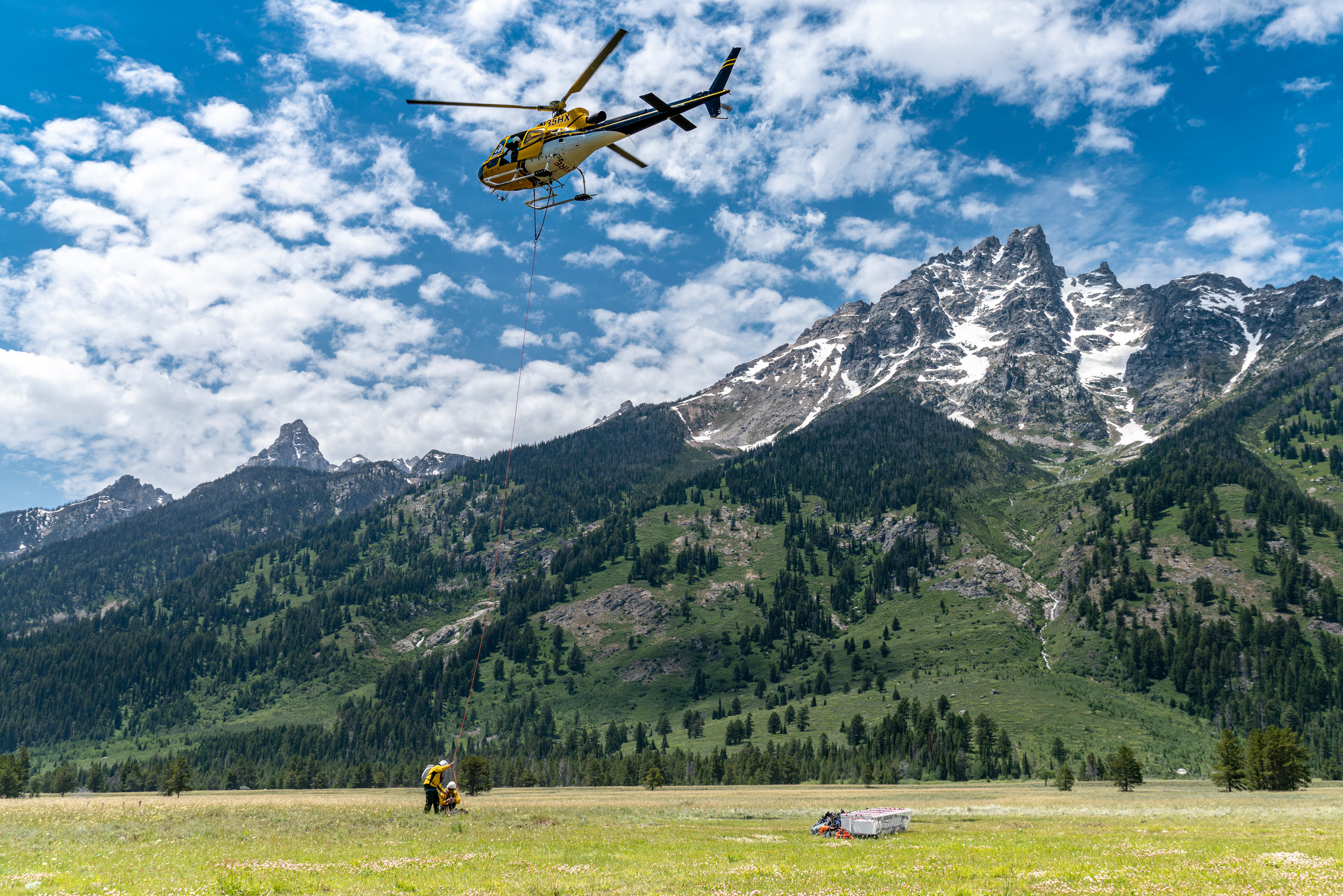 Search and Rescue Two search and rescue personnel stand below helicopter performing emergency response with the Teton range in the background.