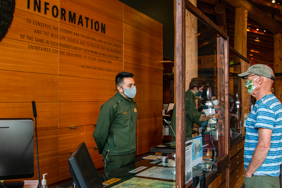 Ranger wearing mask provides information to visitor behind plexiglass window at the Craig Thomas Discovery and Visitor Center desk in Grand Teton