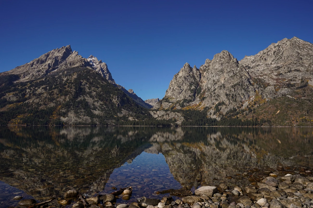 Jenny Lake Trailhead A lake sits at the base of a mountain canyon with a near perfect reflection on its calm surface.