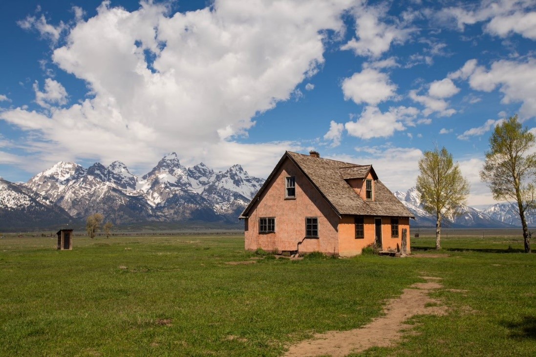 A path leads through grass to a historic pink house with the Teton Range in the background.