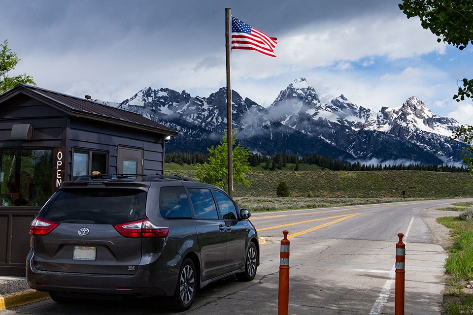 Car at Entrance Station with American Flag on a pole and the Teton Range in the distance