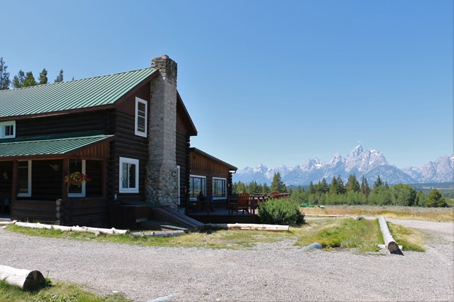 A log building with a large stone chimney and mountains in the background.
