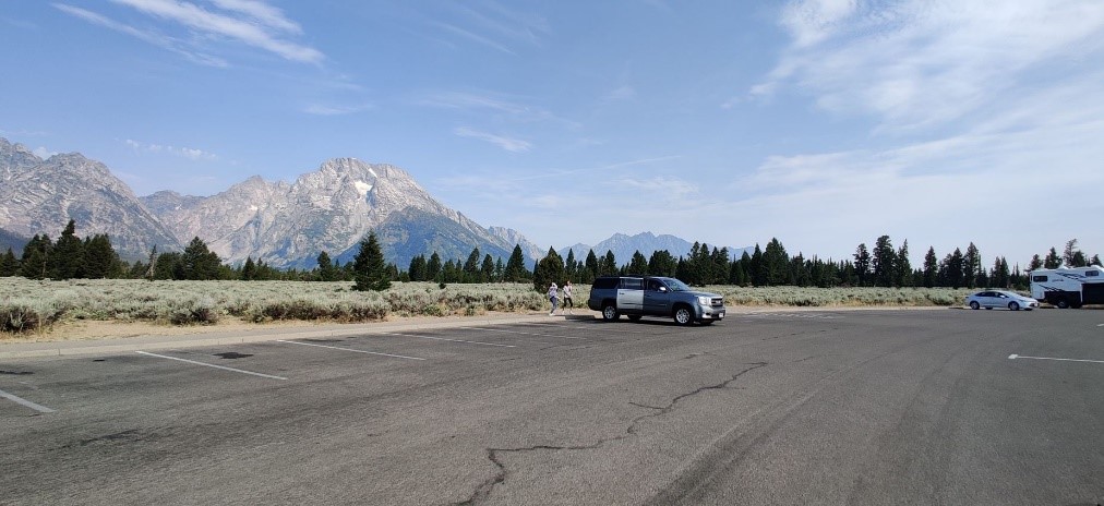 View of a small parking lot with a couple of cars in it and the Teton range in the background. 
