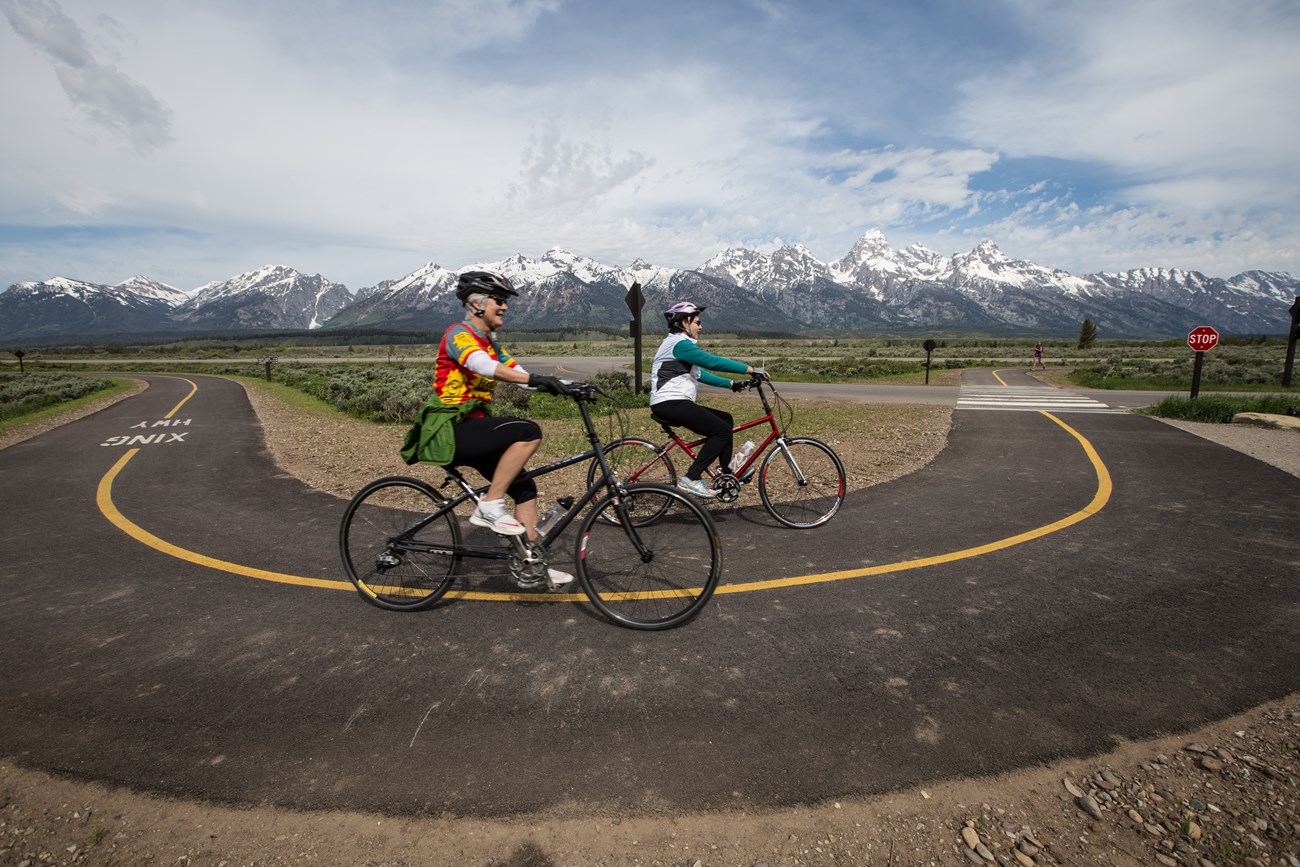 Two bikers ride around a curve on a paved path with mountains in the background.